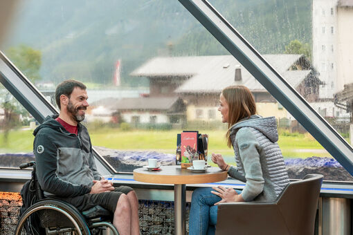 Zwei Freunde bei denen einer im Rollstuhl sitzt unterhalten sich im Cafe Vitalberg mit einem heißen Getränk bei Regenwetter.