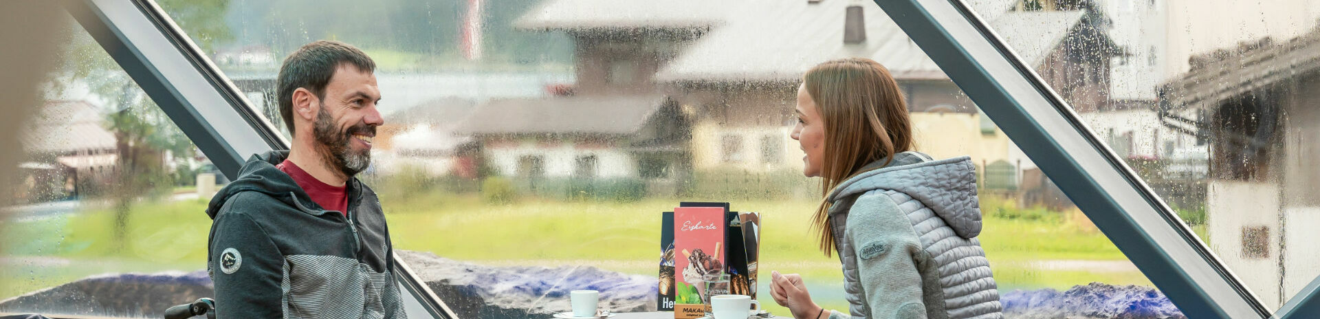 Barrierefreiheit am Achensee Zwei Freunde bei denen einer im Rollstuhl sitzt unterhalten sich im Cafe Vitalberg mit einem heißen Getränk bei Regenwetter.