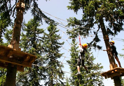 Abenteuerpark Achensee Zwei Kinder mit Kletterausrüstung bewegen sich in einem Hochseilgarten zwischen hohen Nadelbäumen. Sie balancieren auf Seilen und klettern durch ein großes Spinnennetz aus Seilen, das zwischen Holzplattformen in mehreren Metern Höhe gespannt ist. Die Szene zeigt einen sonnigen Tag in der Natur und vermittelt Abenteuer, Bewegung und Geschicklichkeit.