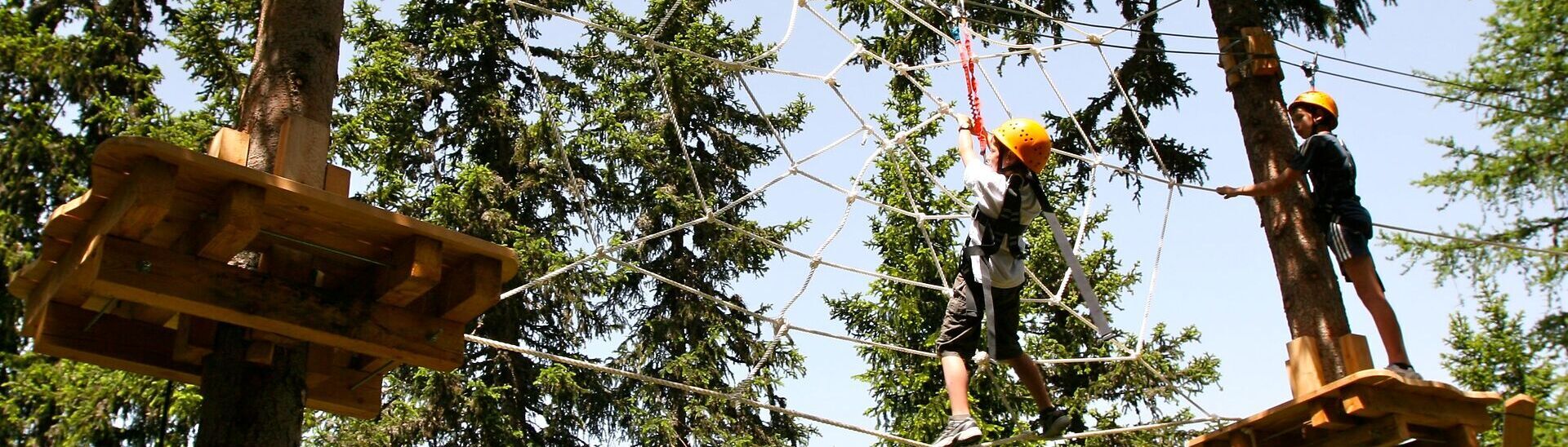 Waldhochseilgarten in Achenkirch am Achensee Zwei Kinder mit Kletterausrüstung bewegen sich in einem Hochseilgarten zwischen hohen Nadelbäumen. Sie balancieren auf Seilen und klettern durch ein großes Spinnennetz aus Seilen, das zwischen Holzplattformen in mehreren Metern Höhe gespannt ist. Die Szene zeigt einen sonnigen Tag in der Natur und vermittelt Abenteuer, Bewegung und Geschicklichkeit.