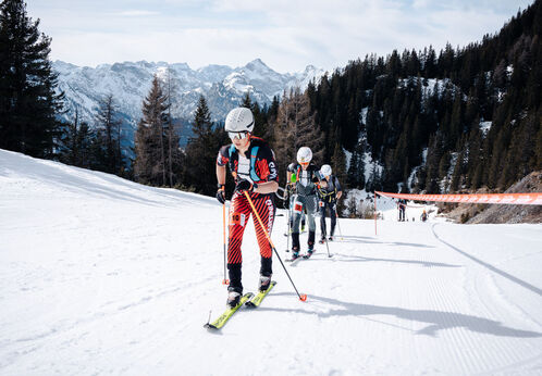 A group of skiers ascends a snowy slope, navigating through the winter landscape. The background features snow-capped mountains and evergreen trees. The skiers are equipped with skiing gear and poles, concentrating on their climb. A red barrier marks the course ahead.