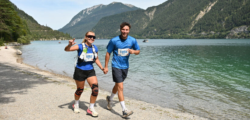 Two runners, a woman and a man, are joyfully holding hands while running along a shoreline. They are wearing matching blue shirts and backpacks, with race numbers visible on their tops. A serene lake and mountains are in the background, creating a picturesque outdoor setting.