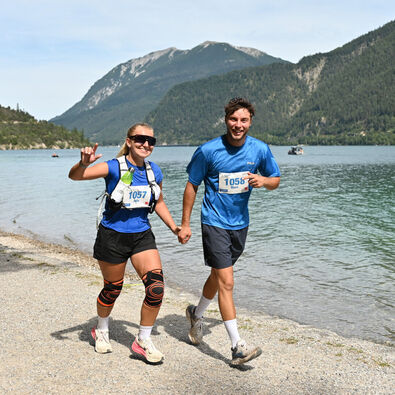 Two runners, a woman and a man, are joyfully holding hands while running along a shoreline. They are wearing matching blue shirts and backpacks, with race numbers visible on their tops. A serene lake and mountains are in the background, creating a picturesque outdoor setting.