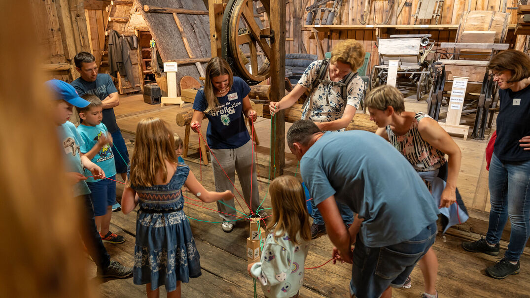 Das Foto zeigt ein Mitmachprogramm in einem historischen Museumsgebäude mit Holzboden und alten landwirtschaftlichen Maschinen. Mehrere Kinder und Erwachsene bilden einen Kreis. Jede Person hält eine rote oder grüne Schnur, die in der Mitte an einem kleinen Holzgestell befestigt ist. Gemeinsam koordinieren sie ihre Bewegungen, um das Objekt zu steuern oder eine Aufgabe zu lösen. Zwei Betreuerinnen stehen im Kreis und unterstützen die Gruppe. Die Teilnehmenden wirken aufmerksam und engagiert. Im Hintergrund befinden sich historische Kutschen, Geräte und Werkzeuge, die den musealen Charakter des Ortes unterstreichen. Die Szene vermittelt Zusammenarbeit, Lernen und gemeinsames Erleben in einer familienfreundlichen Umgebung.