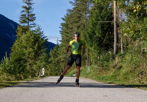 Ein Sportler in einem grünen Trikot fährt auf Skirollern auf einem gepflasterten Weg am Ufer des Achensees entlang.