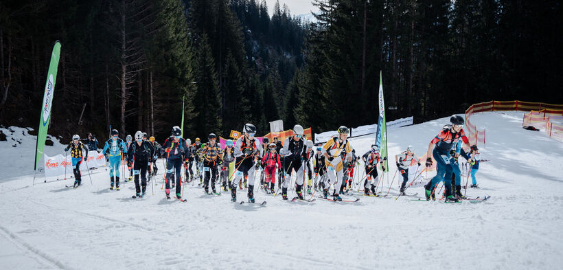 Eine Gruppe von Skifahrern steht auf einer verschneiten Piste im Gebirge, bereit für den Start eines Wettkampfs. Sie tragen Ski- und Sicherheitsausrüstung, umgeben von Bäumen und Banner, die die Veranstaltung kennzeichnen. Der Himmel ist teilweise bewölkt.