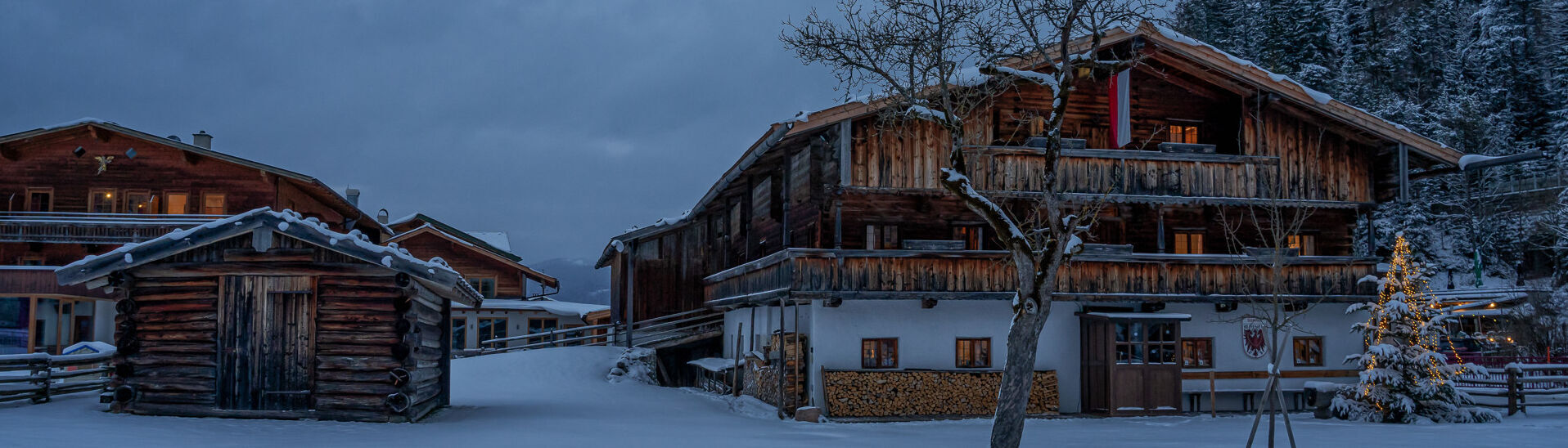 Das Tiroler Heimatmuseum Sixenhof in Achenkirch im Winter. Der Boden ist mit frischem Schnee bedeckt. Im Vordergrund steht ein kleiner Holzschuppen, daneben ein Weihnachtsbaum.