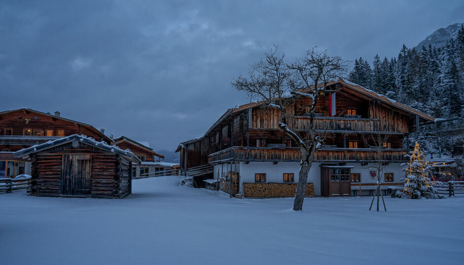 Das Tiroler Heimatmuseum Sixenhof in Achenkirch im Winter. Der Boden ist mit frischem Schnee bedeckt. Im Vordergrund steht ein kleiner Holzschuppen, daneben ein Weihnachtsbaum.