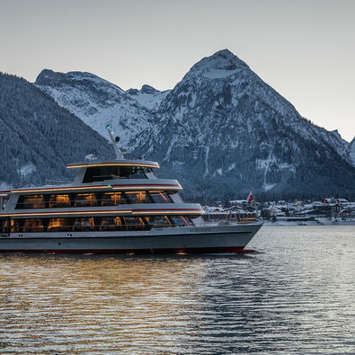 Die MS Achensee bei einer Rundfahrt an einem Winterabend auf dem Achensee. Im Hintergrund sieht man das Dorf Pertisau und das Karwendelgebirge.