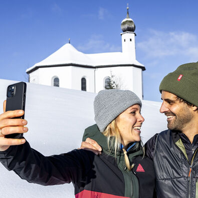 Ein Pärchen möchte den schönen Moment, umgeben von einer herrlichen Winterlandschaft und dem Annakircherl im Hintergrund, mit dem Handy festhalten.