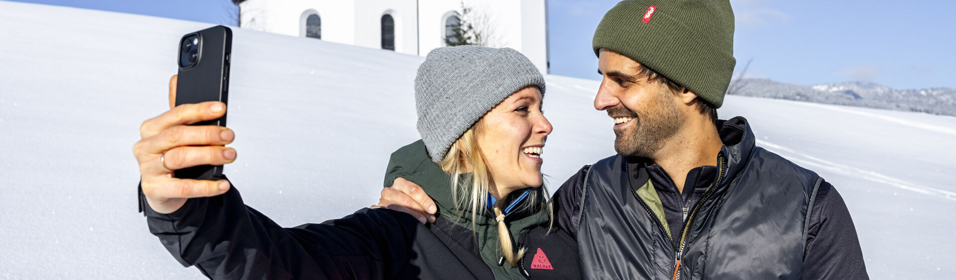 Ein Pärchen möchte den schönen Moment, umgeben von einer herrlichen Winterlandschaft und dem Annakircherl im Hintergrund, mit dem Handy festhalten.