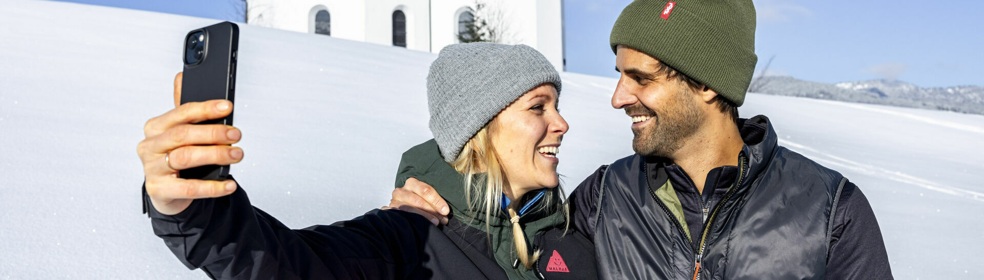 Ein Pärchen möchte den schönen Moment, umgeben von einer herrlichen Winterlandschaft und dem Annakircherl im Hintergrund, mit dem Handy festhalten.