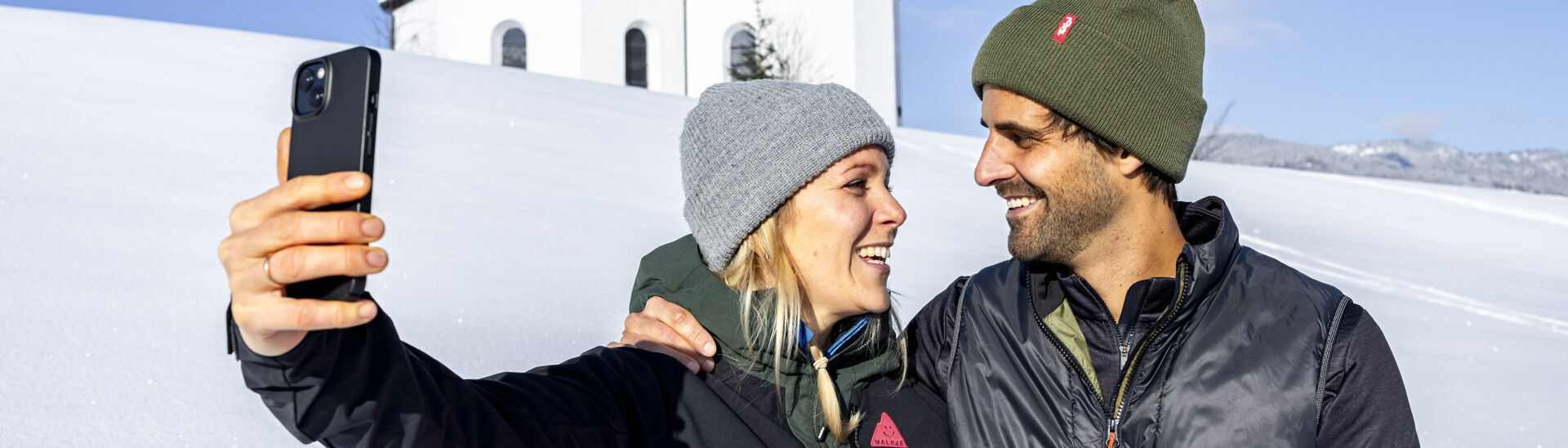 Ein Pärchen möchte den schönen Moment, umgeben von einer herrlichen Winterlandschaft und dem Annakircherl im Hintergrund, mit dem Handy festhalten.