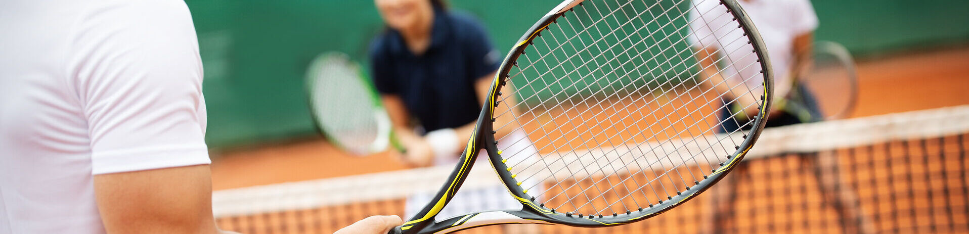 Tennis am Achensee Drei Personen spielen Tennis auf einem Sandplatz. Im Vordergrund hält eine Person einen Tennisschläger bereit, während zwei Mitspieler auf der gegenüberliegenden Seite des Netzes stehen. Die Aufnahme vermittelt die Dynamik und Freude eines Tennismatches im Freien.