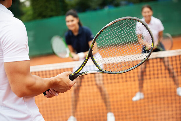 Drei Personen spielen Tennis auf einem Sandplatz. Im Vordergrund hält eine Person einen Tennisschläger bereit, während zwei Mitspieler auf der gegenüberliegenden Seite des Netzes stehen. Die Aufnahme vermittelt die Dynamik und Freude eines Tennismatches im Freien.