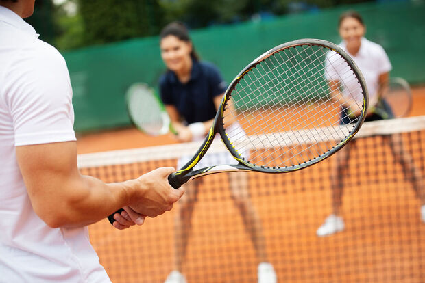 Drei Personen spielen Tennis auf einem Sandplatz. Im Vordergrund hält eine Person einen Tennisschläger bereit, während zwei Mitspieler auf der gegenüberliegenden Seite des Netzes stehen. Die Aufnahme vermittelt die Dynamik und Freude eines Tennismatches im Freien.