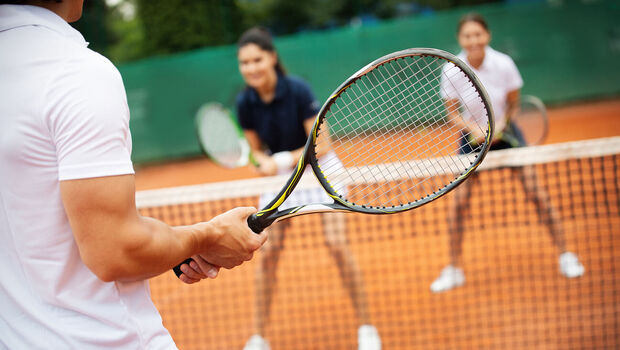 Tennis am Achensee Drei Personen spielen Tennis auf einem Sandplatz. Im Vordergrund hält eine Person einen Tennisschläger bereit, während zwei Mitspieler auf der gegenüberliegenden Seite des Netzes stehen. Die Aufnahme vermittelt die Dynamik und Freude eines Tennismatches im Freien.