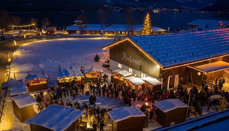 Der Bergadvent beim Fischergut liegt direkt am Ufer des Achensees Bergadvent beim Fischergut am Achensee mit weihnachtlich beleuchteten Holzhütten, zahlreichen Besuchern und verschneiter Winterlandschaft am Achensee.