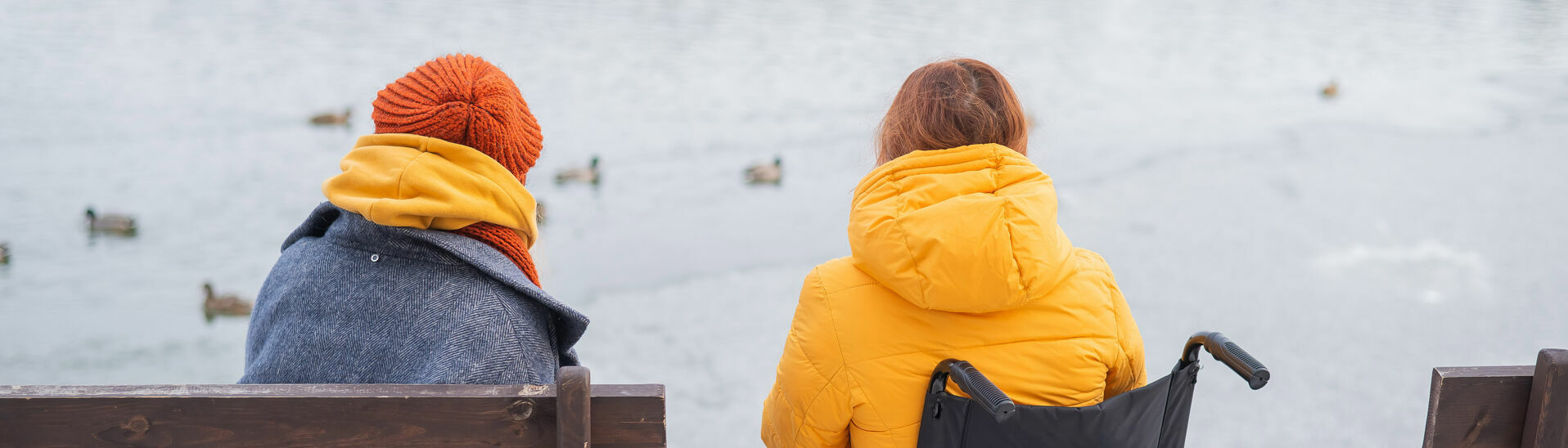 Eine kleine Familie aus einer Rollstuhlfahrerin und einer Frau genießen die Aussicht auf den leicht gefrorener See und dessen winterliche Umgebung.