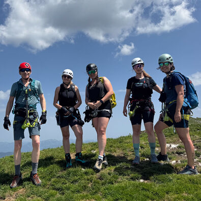 Fünf Personen stehen auf einer Bergwiese. Sie tragen Klettersteigausrüstung. Die Gruppe wirkt entspannt und bereit für das nächste Abenteuer in der Natur.
