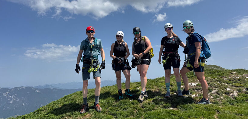 Fünf Personen stehen auf einer Bergwiese. Sie tragen Klettersteigausrüstung. Die Gruppe wirkt entspannt und bereit für das nächste Abenteuer in der Natur.