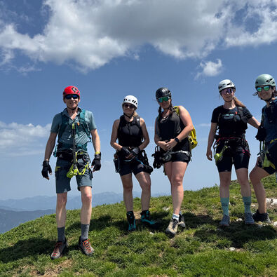 A group of five climbers stands on a grassy peak, wearing helmets and climbing gear. The background features mountains and a clear blue sky with clouds. They appear ready for an outdoor adventure, showcasing a sense of camaraderie and excitement.