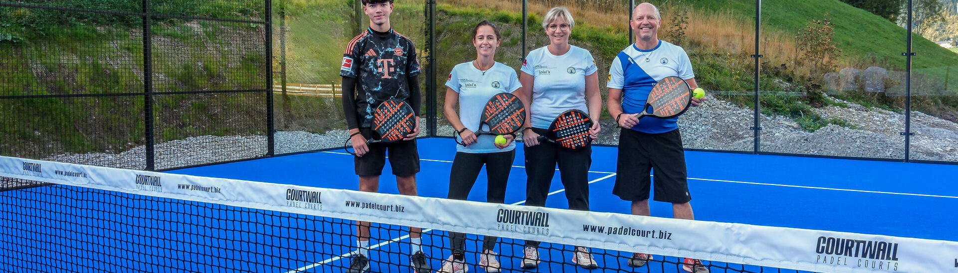 Padelplatz am Achensee Vier Personen stehen mit Padelschlägern auf einem blauen Padelplatz am Achensee in Tirol. Im Vordergrund befindet sich das Netz, im Hintergrund sind die Glaswände des Spielfelds sowie eine grüne Hanglandschaft zu sehen.