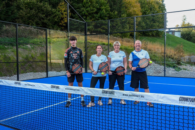 Vier Personen stehen mit Padelschlägern auf einem blauen Padelplatz am Achensee in Tirol. Im Vordergrund befindet sich das Netz, im Hintergrund sind die Glaswände des Spielfelds sowie eine grüne Hanglandschaft zu sehen.