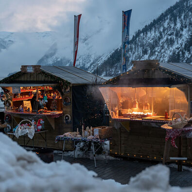 An der Schiffsanlegestelle Scholastika in Achenkirch am Achensee locken hell erleuchtete Marktstände mit Kunsthandwerk zur Landweihnacht. /// The Christmas market on the Scholastika pier in Achenkirch am Achensee attracts visitors with brightly lit market stalls selling artisan wares.