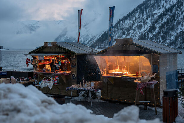 An der Schiffsanlegestelle Scholastika in Achenkirch am Achensee locken hell erleuchtete Marktstände mit Kunsthandwerk zur Landweihnacht. /// The Christmas market on the Scholastika pier in Achenkirch am Achensee attracts visitors with brightly lit market stalls selling artisan wares.