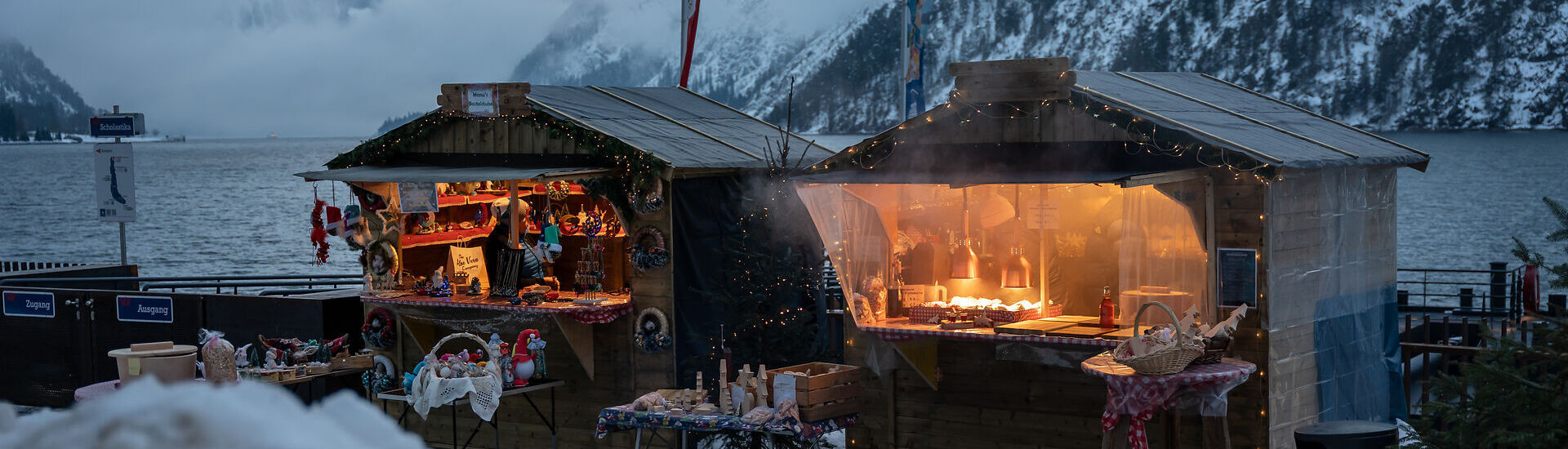An der Schiffsanlegestelle Scholastika in Achenkirch am Achensee locken hell erleuchtete Marktstände mit Kunsthandwerk zur Landweihnacht. /// The Christmas market on the Scholastika pier in Achenkirch am Achensee attracts visitors with brightly lit market stalls selling artisan wares.