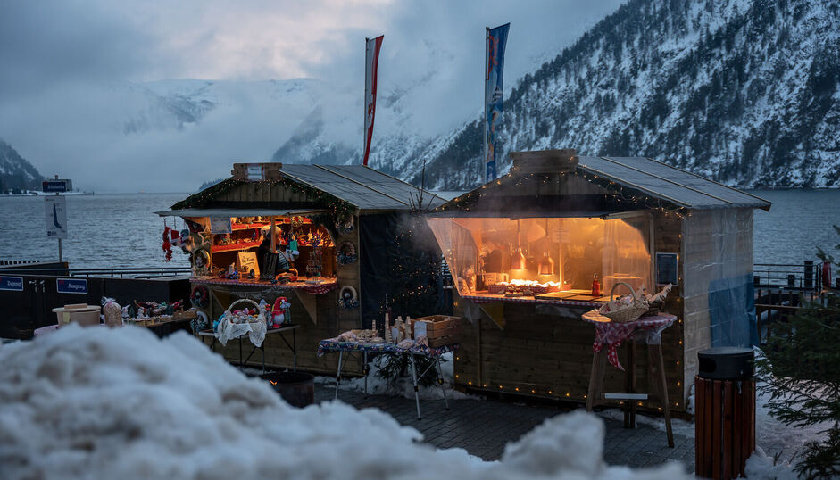 An der Schiffsanlegestelle Scholastika in Achenkirch am Achensee locken hell erleuchtete Marktstände mit Kunsthandwerk zur Landweihnacht. /// The Christmas market on the Scholastika pier in Achenkirch am Achensee attracts visitors with brightly lit market stalls selling artisan wares.