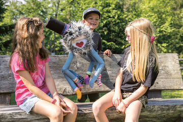 Drei Kinder sitzen auf einer Bank. Ein Junge hält eine farbenfrohe Handpuppe mit einem freundlichen Gesicht und einem fröhlichen Ausdruck. Die Kinder lachen und scheinen Spaß zu haben. Im Hintergrund sind Bäume und grünes Gras zu sehen.