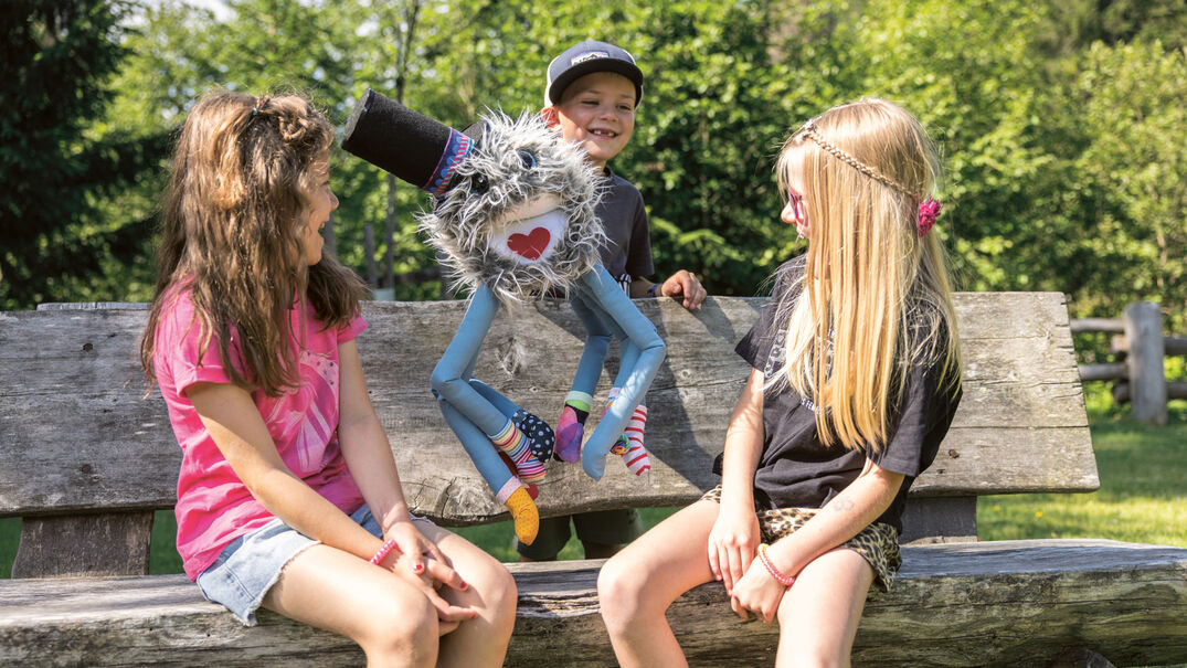Two children sit on a wooden bench, smiling and engaged with a colorful puppet that has playful features and a heart on its face. The scene is set in a bright outdoor environment with greenery in the background.