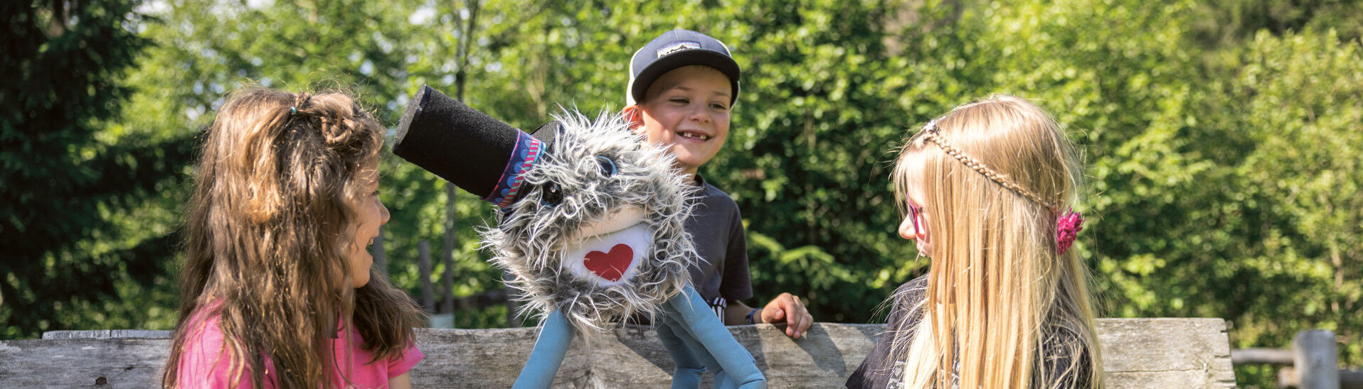 Achensee Kinderprogramm Drei Kinder sitzen auf einer Bank. Ein Junge hält eine farbenfrohe Handpuppe mit einem freundlichen Gesicht und einem fröhlichen Ausdruck. Die Kinder lachen und scheinen Spaß zu haben. Im Hintergrund sind Bäume und grünes Gras zu sehen.