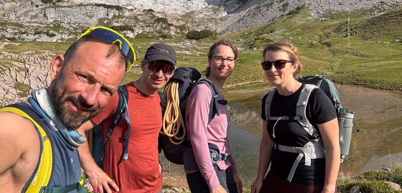 Vier Erwachsene mit Wanderrucksäcken stehen auf einem Bergwanderweg und blicken in die Kamera. Im Hintergrund sind eine alpine Landschaft am Achensee mit felsigen Berghängen und grünen Wiesen zu sehen.