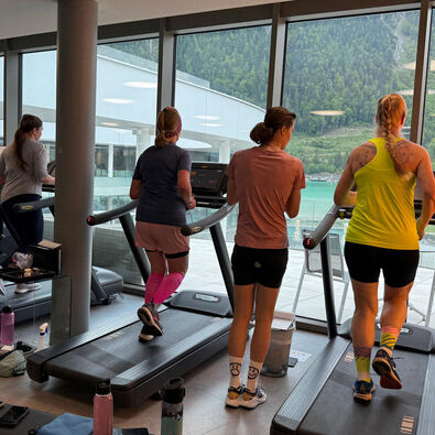 Teilnehmer des Achensee Laufcamps trainiert auf Laufbändern im Lakeside-GYM mit großen Fenstern, die einen Blick auf eine grüne Landschaft und den See bieten. Die Sportler tragen verschiedene Trainingsoutfits und konzentrieren sich auf ihre Aufgaben.