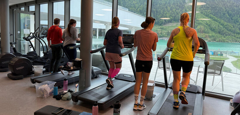 Teilnehmer des Achensee Laufcamps trainiert auf Laufbändern im Lakeside-GYM mit großen Fenstern, die einen Blick auf eine grüne Landschaft und den See bieten. Die Sportler tragen verschiedene Trainingsoutfits und konzentrieren sich auf ihre Aufgaben.