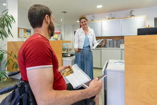 Ein Mann im Rollstuhl spricht mit einer lächelnden Frau im Barrierefreien Infobüro in Pertisau am Achensee.