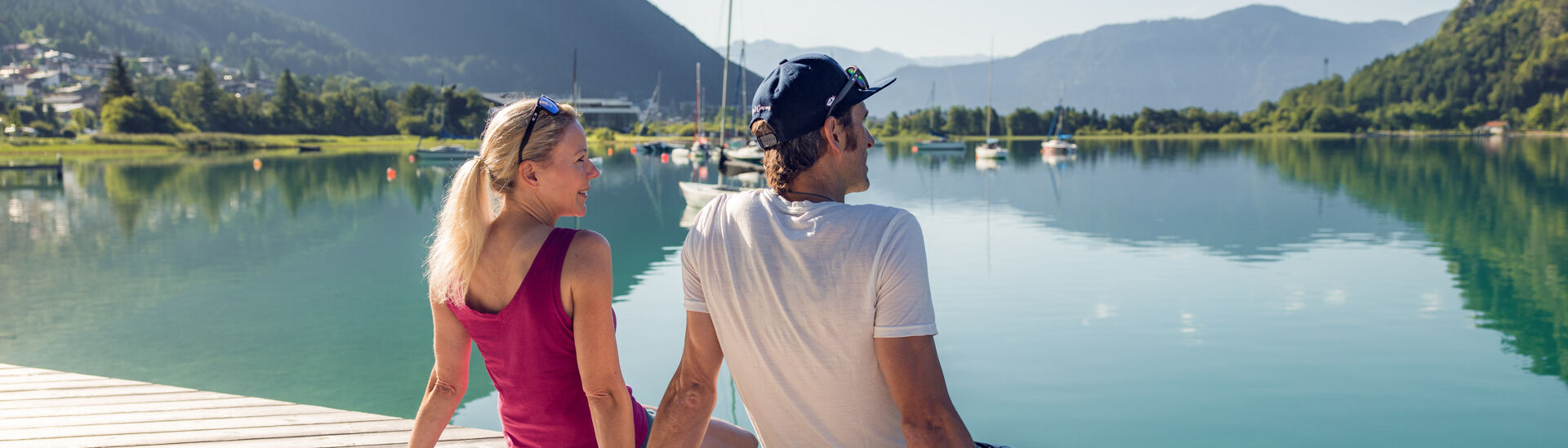 Ein Paar genießt das herrliche Wetter auf dem Holzsteg in Buchau. Im Hintergrund sind Segelboote und das Atoll Achensee zu sehen.