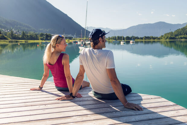 Ein Paar genießt das herrliche Wetter auf dem Holzsteg in Buchau. Im Hintergrund sind Segelboote und das Atoll Achensee zu sehen.