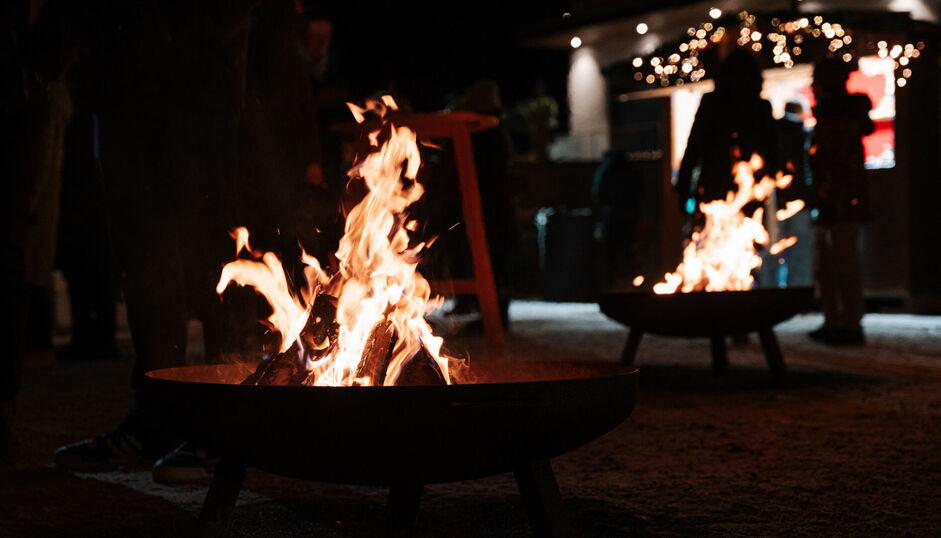 Zahlreiche Feuerschalen sorgen für kuschelige Weihnachtsstimmung Beim Bergadvent in Pertisau brennen zwei Feuerstellen. Im Hintergrund sind Silhouetten von Menschen zu erkennen, die sich um die Feuer versammelt haben.