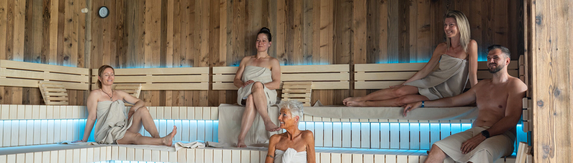 In einer modernen Sauna des Penthouse-SPAs im Atoll Achensee sitzen fünf Menschen in Handtüchern auf Holzbanklinien. Der Raum hat eine warme Atmosphäre mit gedämpftem Licht. In der Mitte ist ein Steingrill sichtbar.