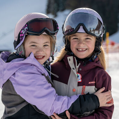 Zwei lächelnde Mädchen stehen im Schnee, die sich umarmen. Beide tragen Skihelme und Brillen. Im Hintergrund sind verschneite Berge und eine Skipiste am Achensee zu sehen, die Atmosphäre wirkt fröhlich und winterlich.