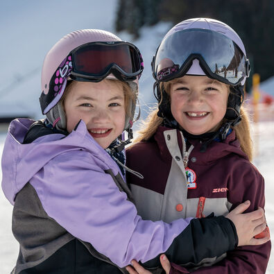 Two young girls are smiling and hugging, wearing ski helmets and colorful winter jackets. They appear happy and playful, standing on a snowy landscape at Lake Achensee with ski slopes in the background. The atmosphere conveys joy and friendship in a winter sports setting.