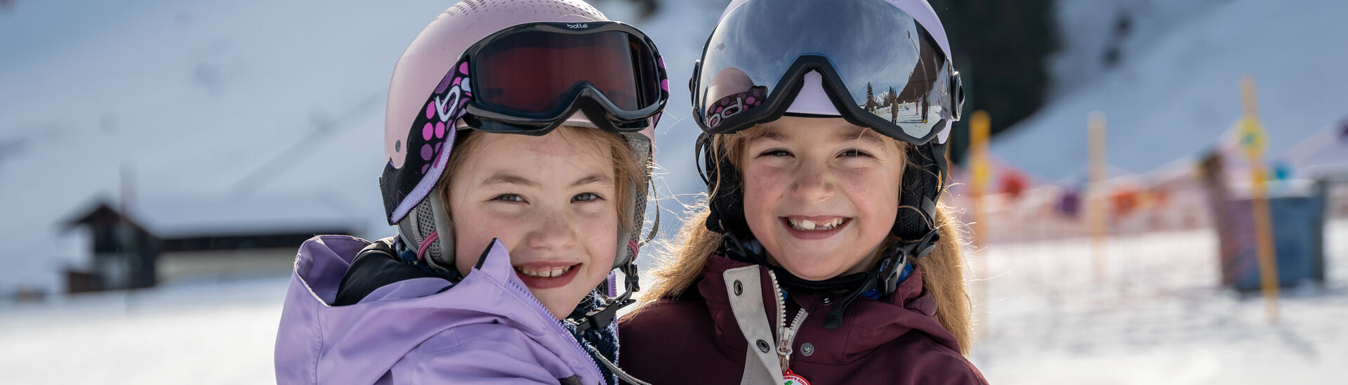 Skifahren bei den Planberg- & Wiesenliften Zwei lächelnde Mädchen stehen im Schnee, die sich umarmen. Beide tragen Skihelme und Brillen. Im Hintergrund sind verschneite Berge und eine Skipiste am Achensee zu sehen, die Atmosphäre wirkt fröhlich und winterlich.