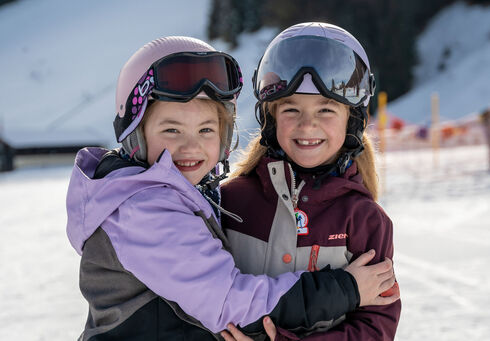 Zwei lächelnde Mädchen stehen im Schnee, die sich umarmen. Beide tragen Skihelme und Brillen. Im Hintergrund sind verschneite Berge und eine Skipiste am Achensee zu sehen, die Atmosphäre wirkt fröhlich und winterlich.