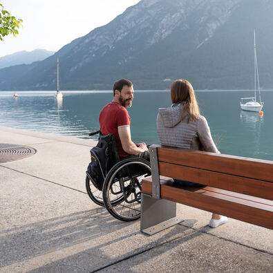  Ein Rollstuhlfahrer und eine Frau sitzen an der Seepromenade in Pertisau im Sommer und schauen auf den See.
