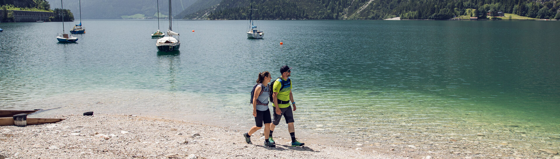 Ein Paar genießt einen kleinen Spaziergang am Ufer des Achensees in Achenkirch. Im Hintergrund sieht man Segelboote.