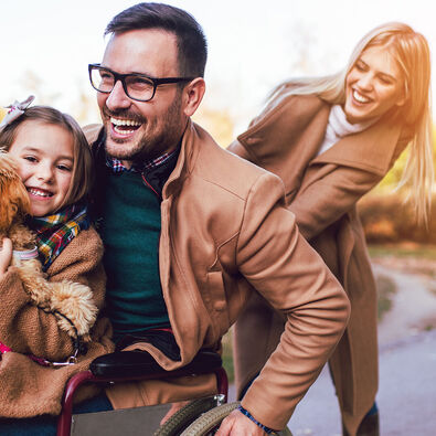Eine lachende Familie aus einem Rollstuhlfahrer, seiner Frau, Tochter und Hund im Sommer.